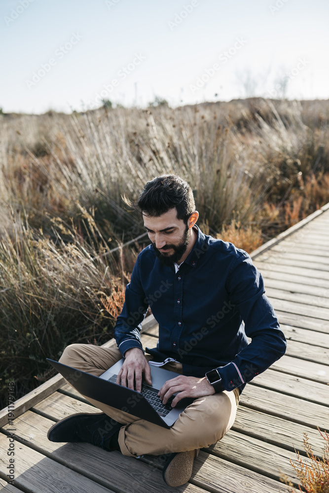 Inspired man working with laptop in natureCasual adult man enjoying freedom in sunny field using laptop while sitting on wooden pier. Inspired man working with laptop in natureCasual adult man enjoying freedom in sunny field using laptop while sitting on wooden pier.