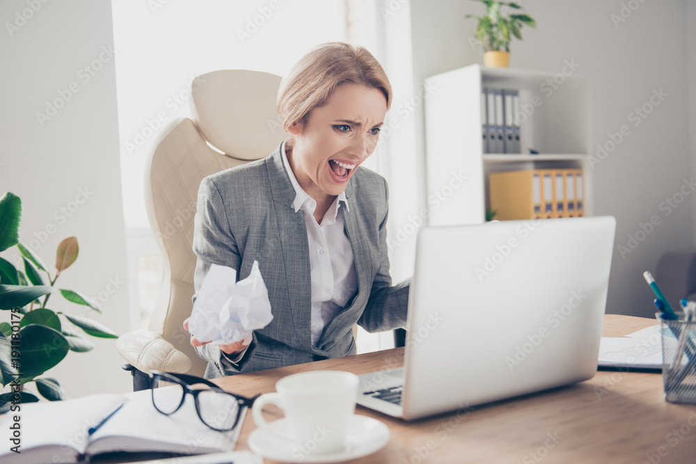 Angry, disappointed, surprised woman in jacket holding crumpled paper in hand, shouting, creaming, looking at laptop, sitting on armchair at her desktop Angry, disappointed, surprised woman in jacket holding crumpled paper in hand, shouting, creaming, looking at laptop, sitting on armchair at her desktop