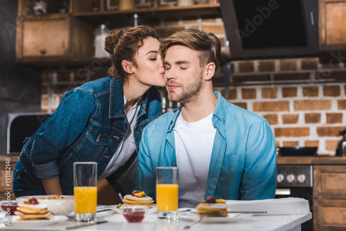 girl kissing boyfriend sitting at table with juice in glasses and pancakes