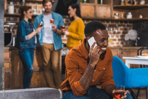 happy young african american man talking by smartphone while friends drinking wine behind