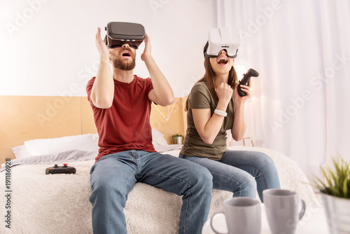 Favorite game. Young pretty pleasant couple relaxing in bedroom while playing in VR glasses and opening mouths