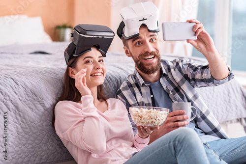 One more shot. Awesome loving gay couple in VR glasses chortling while taking selfie and enjoying popcorn and coffee photo