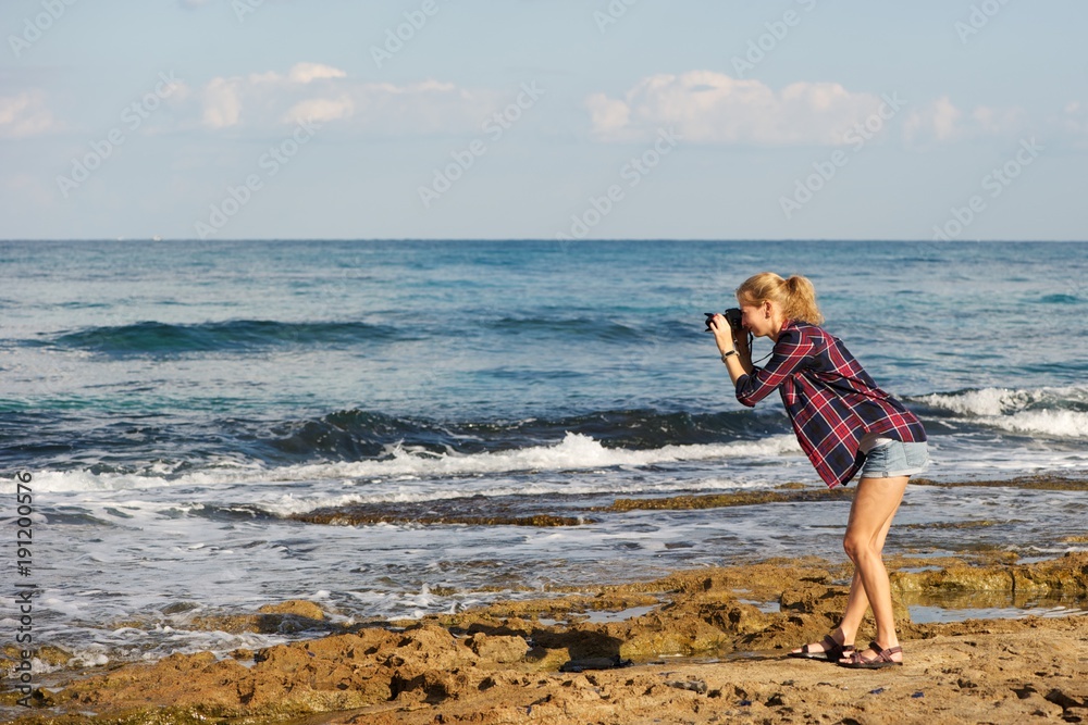 A young woman taking pictures at a beach; wearing shorts and plaid shirt; blonde, pony tail. Standing on sea rocks; sea, sky, clouds in background.