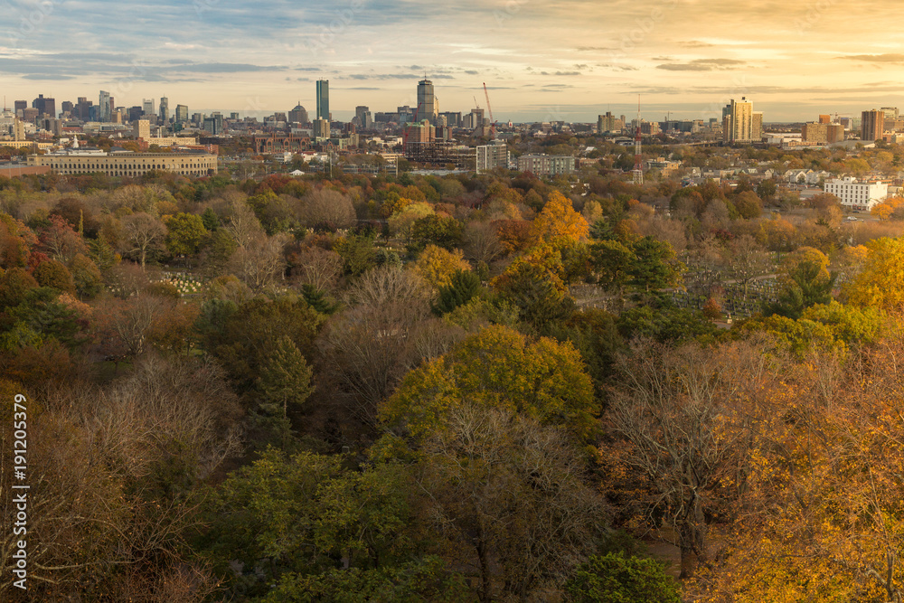 A distant view of a sunset over Boston through a forest of colorful autumn trees. 