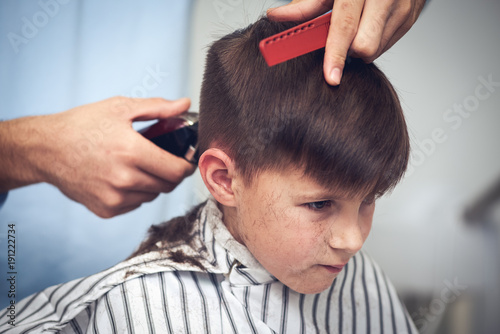 Barber is doing haircut to Caucasian boy using hair clipper in barbershop.