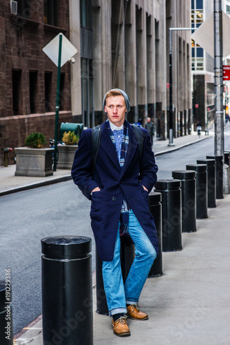 Slika na platnu American man traveling in New York, wearing blue long overcoat, scarf, jeans, leather shoes, cuffed knit  hat, carrying back bag, hands in pockets, sitting on metal pillar on vintage street, relaxing
