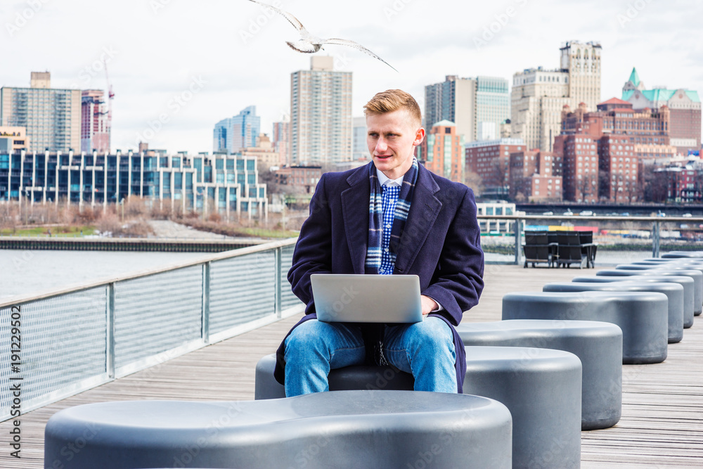 Young American College Student traveling, studying in New York, wearing blue long woolen overcoat, scarf, jeans, sitting by East River, working on laptop computer. Brooklyn buildings on background. Young American College Student traveling, studying in New York, wearing blue long woolen overcoat, scarf, jeans, sitting by East River, working on laptop computer. Brooklyn buildings on background.