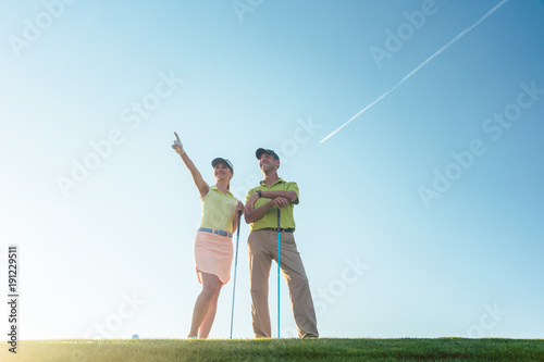 Low-angle view of the silhouette of a man pointing to the horizon, while standing next to his female partner on a professional golf course against sunshine and clear blue sky