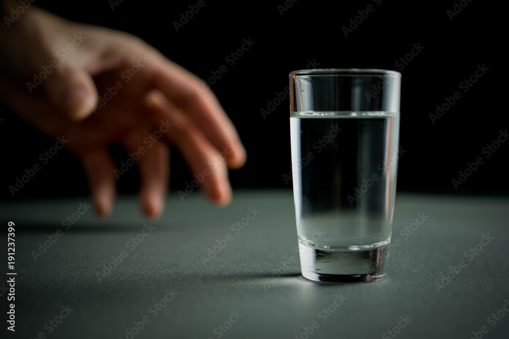 hand reaches for a glass of vodka or alcohol drink, alcoholism and alcohol abuse concept, defocused,.selective focus, close up, gray table, dark background hand reaches for a glass of vodka or alcohol drink, alcoholism and alcohol abuse concept, defocused,.selective focus, close up, gray table, dark background
