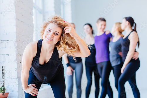 Close up image of attractive curly blond fitness woman over blurred group of people in white gym interior. Teamwork, good mood and healthy lifestyle concept. © Iryna