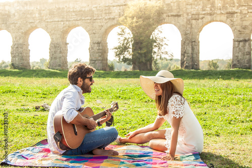 Beautiful girl with hat with young man playing guitar in park on a sunny day. Happy couple sitting on colorful picnic blanket in front of roman aqueduct ruins in rome.