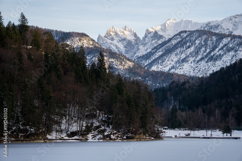 Fototapeta Naklejka Na Ścianę i Meble -  Frozen lake in the foreground with rugged alpine peaks in the background. The late evening rays of sun are shining on the snowcapped mountains
