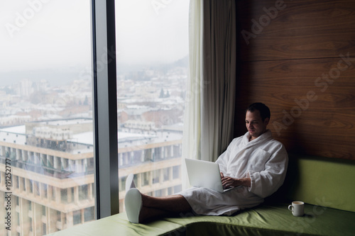 Businessperson working at home or in trip. Side view portrait of handsome young businessman sitting on bed and using laptop. Panoramic window with beautiful dawn city scenery on the background