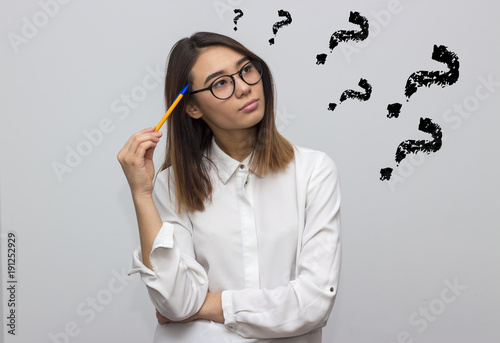 Indoor portrait of beautiful brunette asian woman with pencil and glasses having a lot of questions. Young student or businesswoman concept. Selective focus and shallow DOF