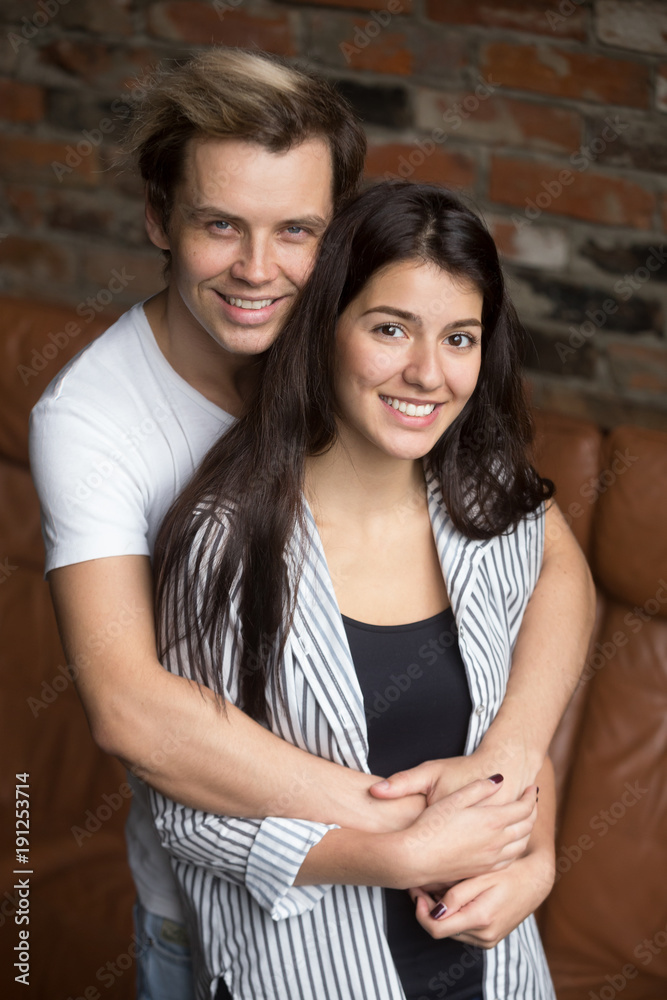 Vertical portrait of smiling attractive young couple looking at camera, cheerful millennial casual man and woman embracing at home, happy teen girl and guy dating hugging standing together indoors