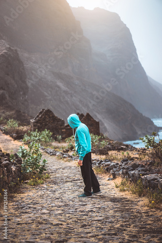 Tourist in hoodie in Aranhas valley hiking to Ponta do Sol. Huge mountains of coastline and old local stone house in the background. Santo Antao Island, Cape Verde photo