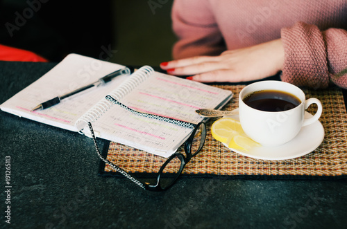 The glasses, notebook and hot tea on table with famale hands, start up time concept.