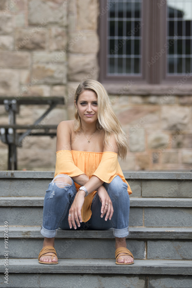 Stunning young blonde fashion model poses in orange blouse with bare shoulders and blue jeans - sitting on front porch steps