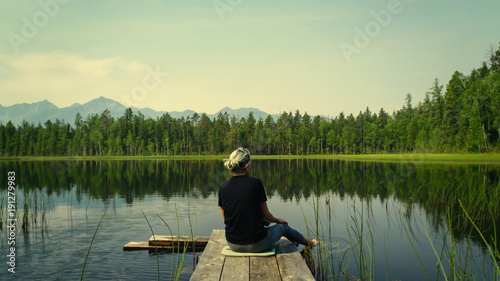 Lonely dreaming woman sitting on a wooden bridge pier looking at the mountains reflected in the mirror water of the forest lake. Adventure nature concept. Absolute serenity