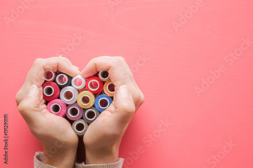 Colorful spools of threads in hands heart shape on the pastel pink background. Womanly hobby. Sewing concept. Empty place for text.
