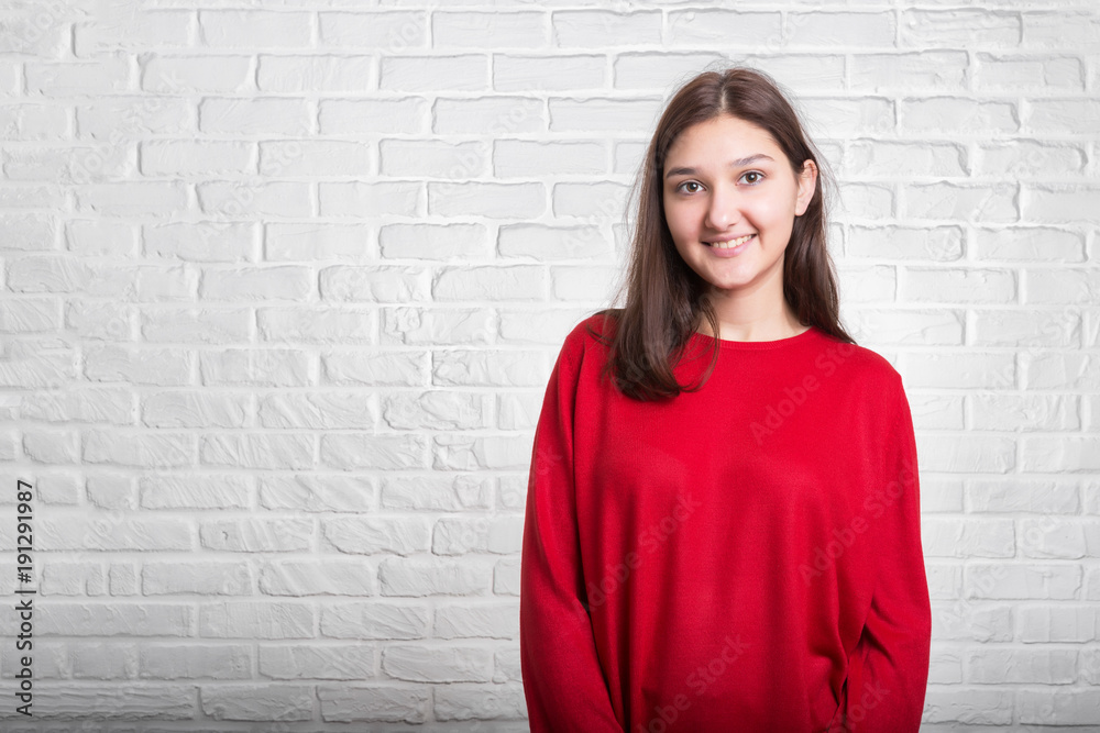 A young pretty smiling girl in a red sweater and a hat against a modern white brick wall in the loft style. She looks at the viewer. Copy space. A young pretty smiling girl in a red sweater and a hat against a modern white brick wall in the loft style. She looks at the viewer. Copy space.