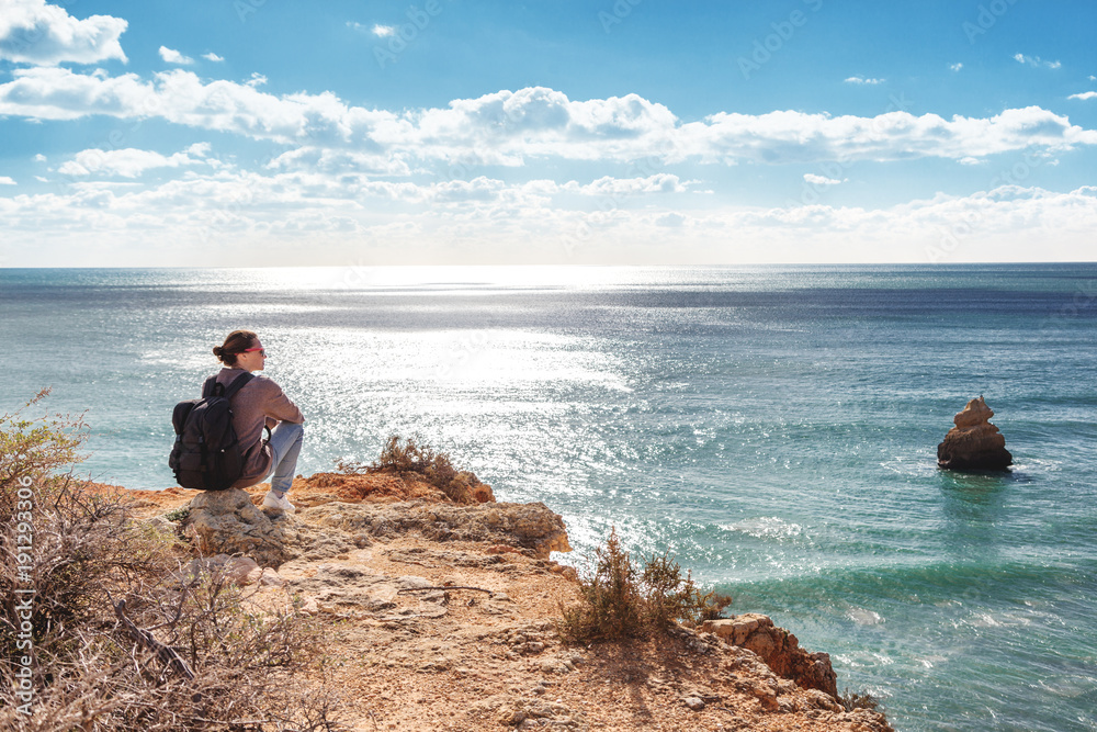 Girl traveler with a backpack sits on the rocks on the ocean, admiring the incredible scenery. Portugal, the Algarve, a popular destination for travel in Europe