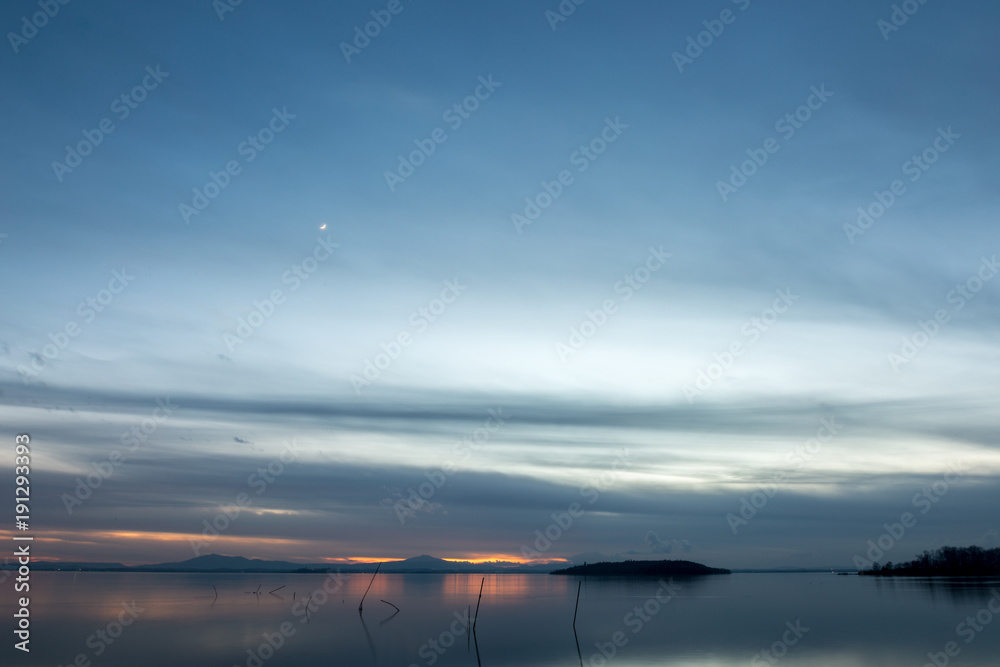 Beautiful view of Trasimeno lake (Umbria, Italy) at dusk, with blue and orange tones and moon in the sky