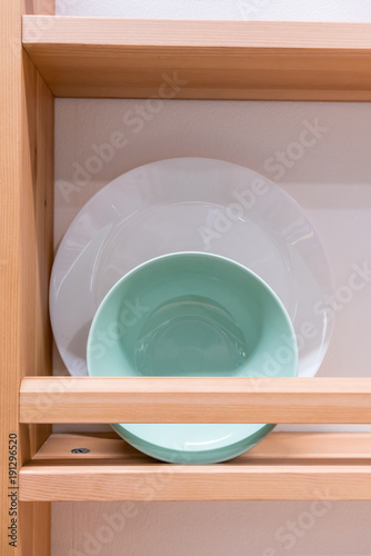 Green ceramic bowl and white porcelain plate on wooden rack against white wall.