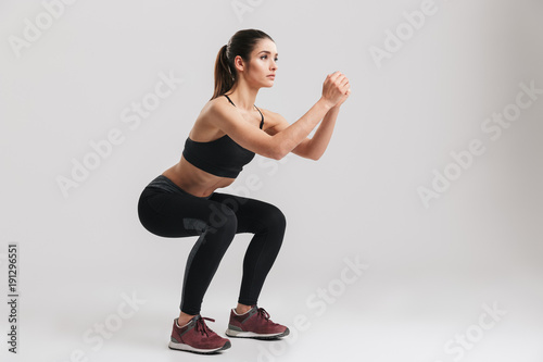 Image of sporty athletic woman in sneakers and tracksuit squatting doing sit-ups in gym, isolated over gray background photo