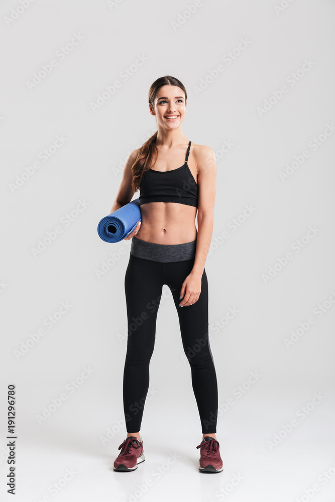 Full-length photo of healthy young woman in sportswear holding yoga mat while practising in studio, isolated over gray background Full-length photo of healthy young woman in sportswear holding yoga mat while practising in studio, isolated over gray background