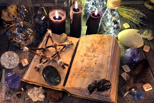 Old witch book with pentagram, black candles, crystals and ritual objects. Occult, esoteric, divination and wicca concept. No foreign text, all symbols on pages are fantasy, imaginary ones 