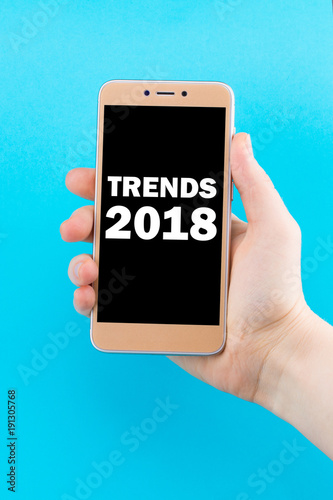 Trends 2018 inscription on phone screen. Golden phone in hand on a blue background photo