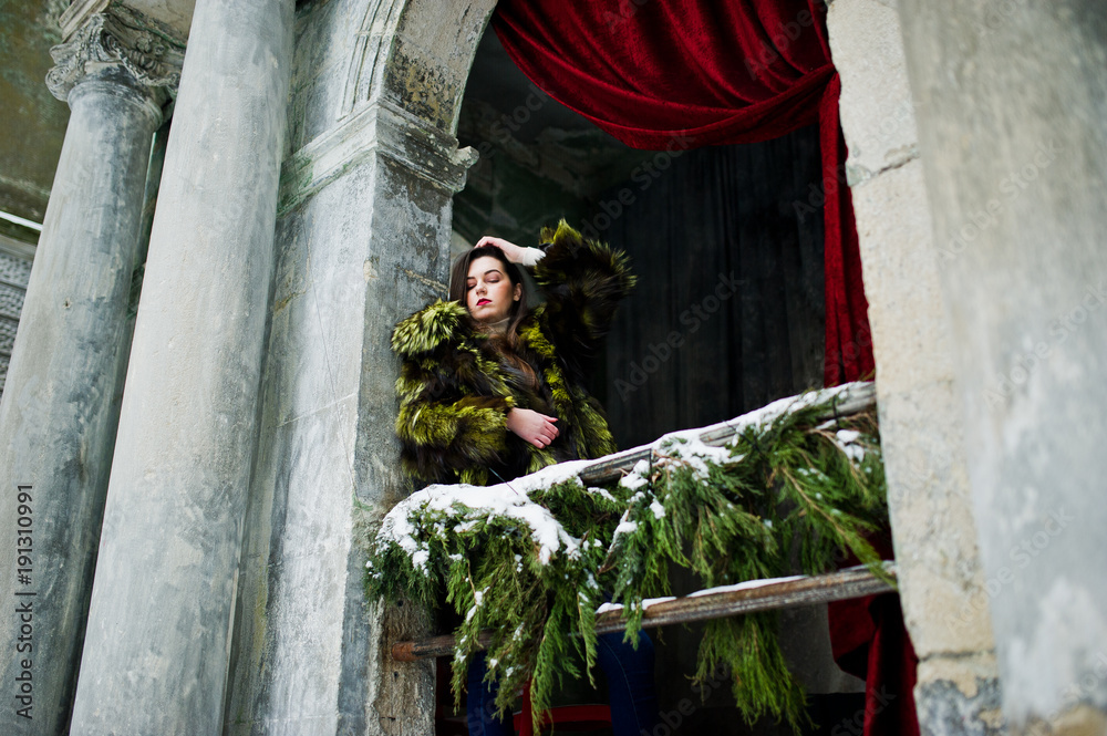 Brunette girl in green fur coat against old arch with columns and red curtains.