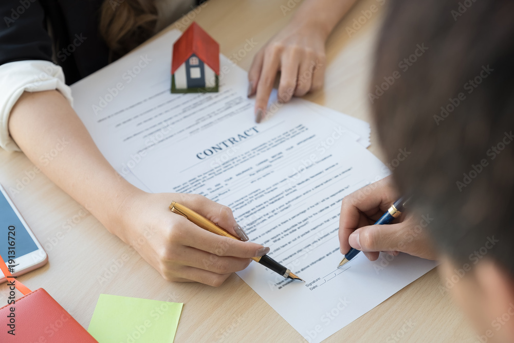 Cropped image of real estate agent assisting client to sign contract paper at desk with house model Cropped image of real estate agent assisting client to sign contract paper at desk with house model
