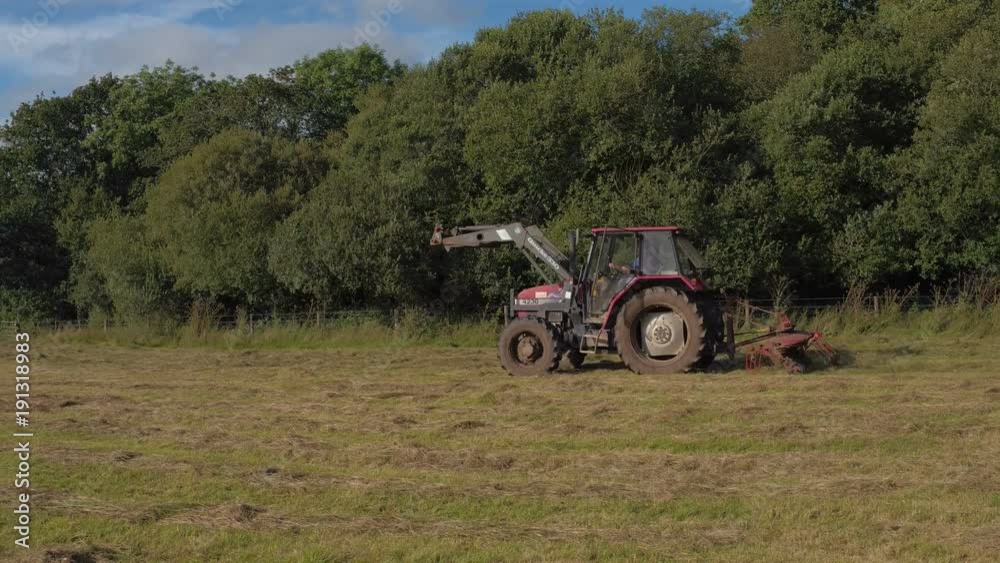 Devon, United Kingdom. 2017 July. Side panning shot of a red tractor clearing grass on a field Devon, United Kingdom. 2017 July. Side panning shot of a red tractor clearing grass on a field