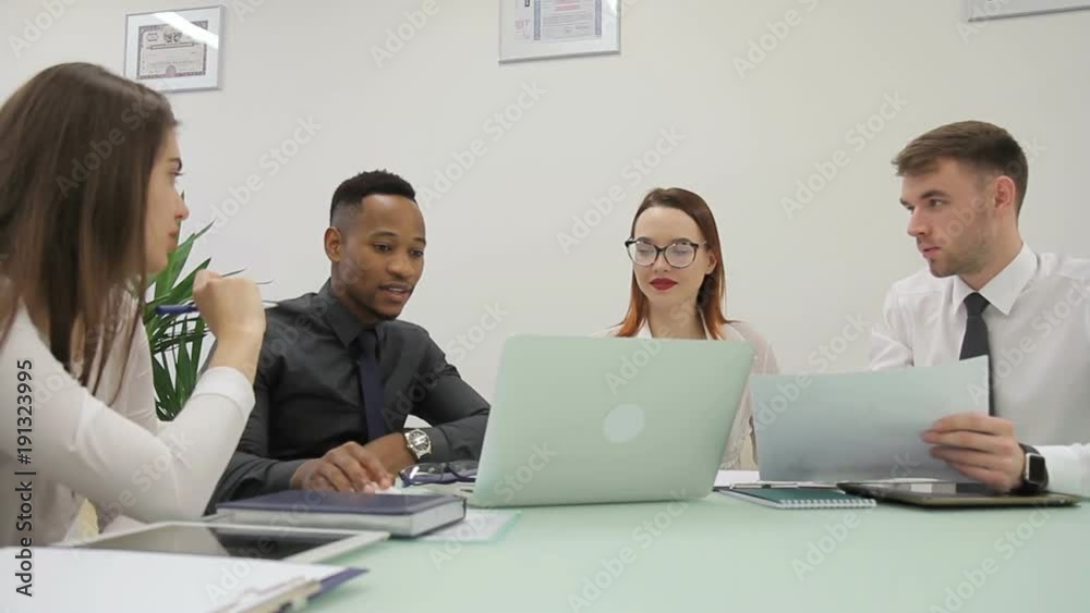 African manager is presenting the results of his report to his colleagues in co-working room. Multiethnical businesspeople are sitting at the table with laptop and tablets on and caucasian businessman African manager is presenting the results of his report to his colleagues in co-working room. Multiethnical businesspeople are sitting at the table with laptop and tablets on and caucasian businessman