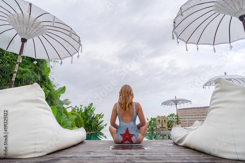 A young girl in a swimsuit is sitting in a yoga pose near the pool, in the background there are greens and palms. Luxury hotel with bungalow. Everything is white and bright. View with copy space