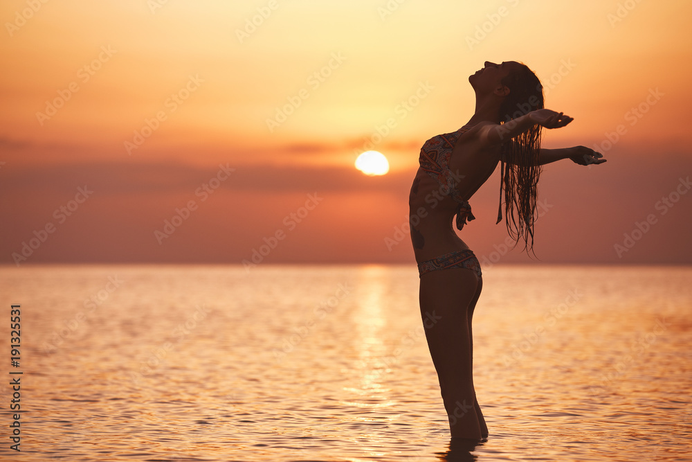 silhouette of a beautiful sexual girl against the sea and sunset. happy woman stands in the sea with wide opened hands and enjoys a summer vacation
