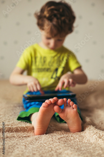 Kid with tablet sitting on bed and watching cartoons. Selective Focus on the feet. Cute boy in green t-shirt and blue pants. Vertical photo