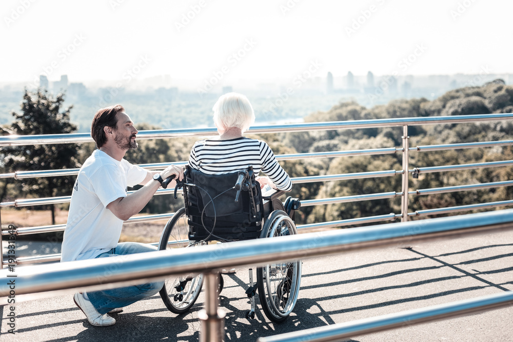 Care and support. Handsome nice bearded man holding a wheelchair and looking at the elderly woman while caring about her