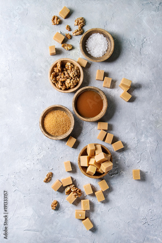 Salted caramel fudge candy served with fleur de sel, caramel sauce, brown cane sugar and caramelized walnuts in wooden bowls over grey texture background. Top view, space. Dessert set