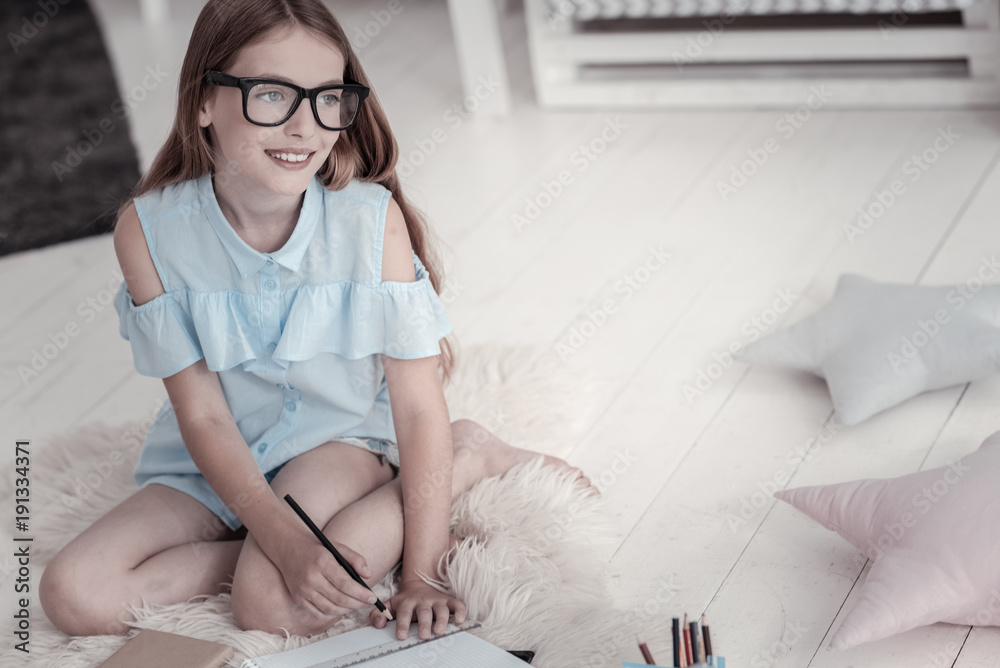 Enjoying free time. Good-looking content smiling girl wearing a blue dress and glasses and writing in her notebook while sitting on the floor and cushions hear her Enjoying free time. Good-looking content smiling girl wearing a blue dress and glasses and writing in her notebook while sitting on the floor and cushions hear her