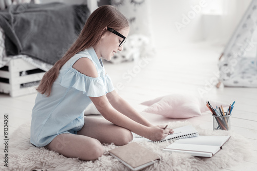 Doing homework. Good-looking cheerful smiling girl wearing a blue dress and glasses and writing in her notebook while sitting on the floor with her books near her