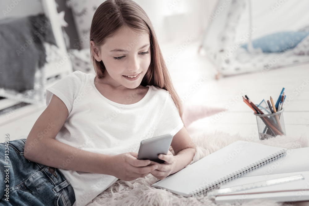 Sending messages. Attractive vigorous smiling long-haired girl wearing jeans and a white t-shirt and holding her black phone while lying on the floor and her stuff near her Sending messages. Attractive vigorous smiling long-haired girl wearing jeans and a white t-shirt and holding her black phone while lying on the floor and her stuff near her