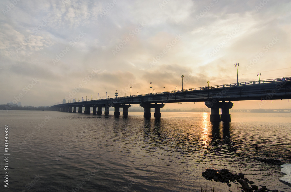 Picturesque view over the Paton's Bridge over the Dnieper River during winter sunrise. Sun reflected in the water. Kyiv, Ukraine. Selective focus with wide angle lens