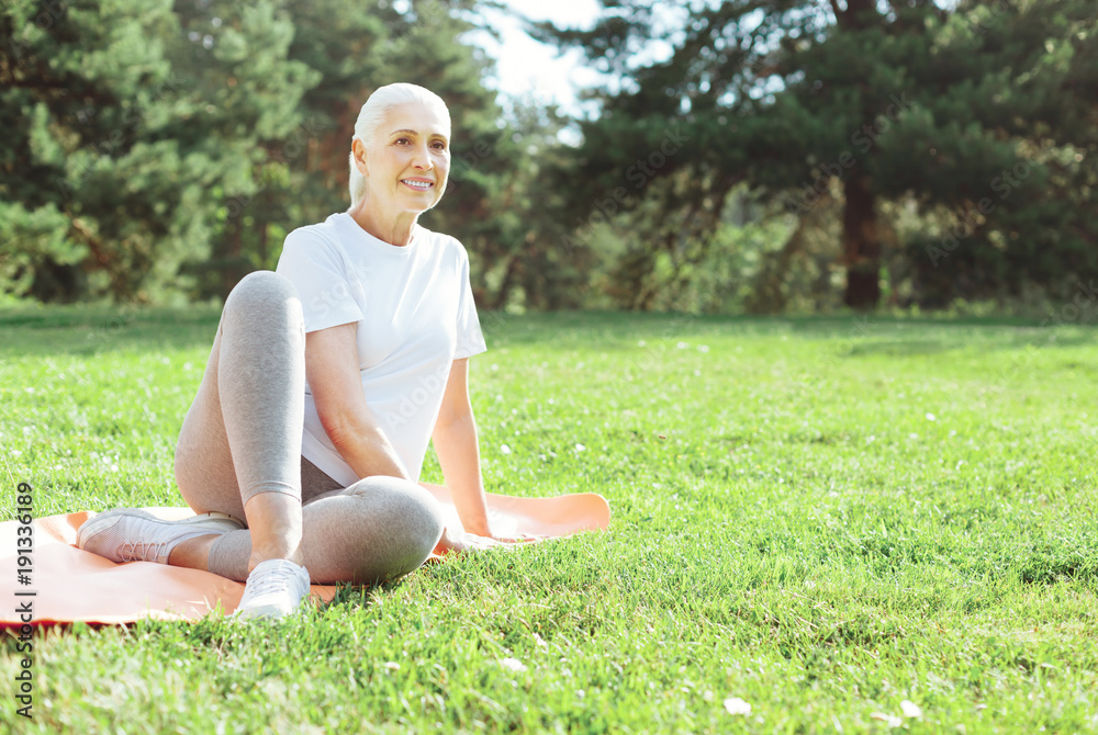 Outdoor exercise. Joyful positive elderly woman sitting on a yoga amt and smiling while exercising outdoors Outdoor exercise. Joyful positive elderly woman sitting on a yoga amt and smiling while exercising outdoors