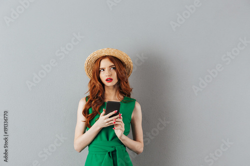 Portrait of thoughtful ginger woman wearing straw hat and green dress looking aside with smartphone in hand, isolated over gray background © Drobot Dean