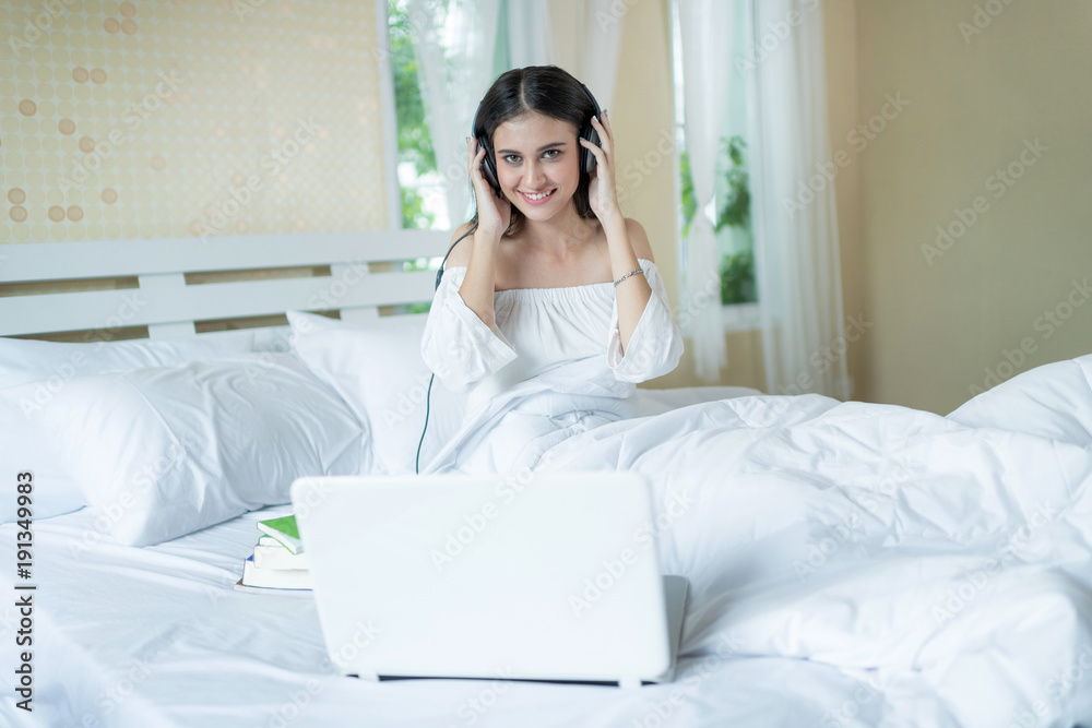 Charming woman on bed wearing headphone enjoy music and using laptop computer .