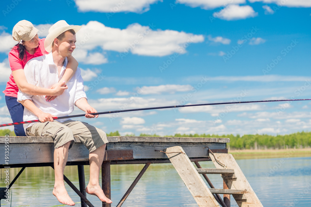 on a wooden pier, a loving couple with a fishing rod is fishing in the lake on a sunny day