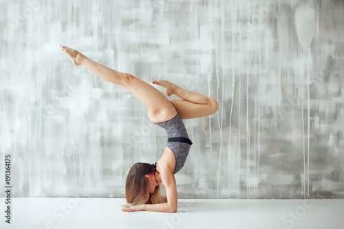 Fototapet Young skinny woman in grey leotard doing yoga handstand in light grey studio